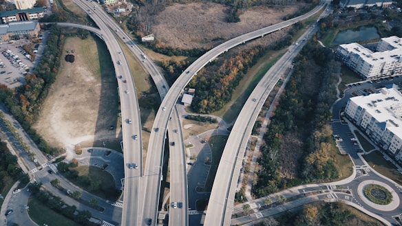 An aerial view of a complex highway interchange with several elevated roads crossing each other. Vehicles are seen traveling in both directions, surrounded by green spaces, urban buildings, and parking lots. Trees and vegetation line the roads, suggesting a mix of urban and natural environments.