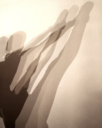 Multiple elongated shadows of a person in a dynamic stretching pose appear on a plain background. The shadows overlap and create a layered effect, suggesting movement and depth.
