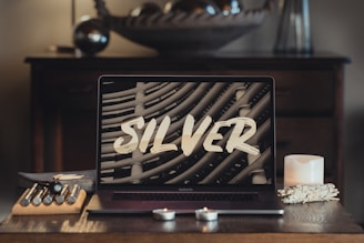 A modern laptop with the word 'SILVER' displayed on its screen sits on a wooden table. The surrounding items include candles, artist brushes in a holder, and decorative elements creating a cozy and artistic atmosphere. In the background, an out-of-focus piece of decor and wooden drawers enhance the scene.