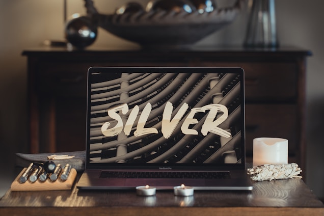 A modern laptop with the word 'SILVER' displayed on its screen sits on a wooden table. The surrounding items include candles, artist brushes in a holder, and decorative elements creating a cozy and artistic atmosphere. In the background, an out-of-focus piece of decor and wooden drawers enhance the scene.