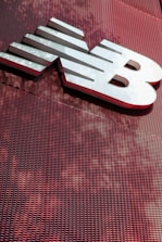 A large, metallic logo is mounted on an industrial-style red mesh background. The logo consists of bold, white letters with a sleek and modern design.