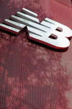 A large, metallic logo is mounted on an industrial-style red mesh background. The logo consists of bold, white letters with a sleek and modern design.