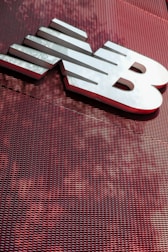 A large, metallic logo is mounted on an industrial-style red mesh background. The logo consists of bold, white letters with a sleek and modern design.