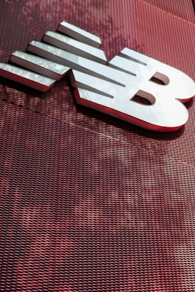 A large, metallic logo is mounted on an industrial-style red mesh background. The logo consists of bold, white letters with a sleek and modern design.