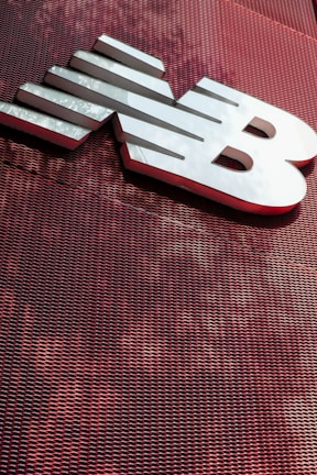 A large, metallic logo is mounted on an industrial-style red mesh background. The logo consists of bold, white letters with a sleek and modern design.