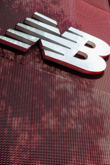 A large, metallic logo is mounted on an industrial-style red mesh background. The logo consists of bold, white letters with a sleek and modern design.