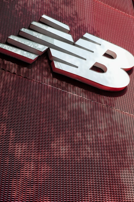 A large, metallic logo is mounted on an industrial-style red mesh background. The logo consists of bold, white letters with a sleek and modern design.