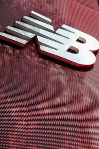 A large, metallic logo is mounted on an industrial-style red mesh background. The logo consists of bold, white letters with a sleek and modern design.