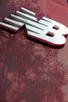 A large, metallic logo is mounted on an industrial-style red mesh background. The logo consists of bold, white letters with a sleek and modern design.