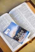 An open book with text in the English language, possibly a religious scripture, has a booklet placed on top of it. The booklet has an image of a scenic landscape and the words 'Talking with God' printed on the cover.