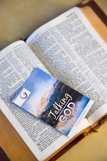 An open book with text in the English language, possibly a religious scripture, has a booklet placed on top of it. The booklet has an image of a scenic landscape and the words 'Talking with God' printed on the cover.