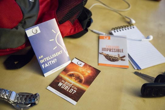 A collection of items is arranged on a light-colored surface, including a red and gray backpack, a blue pamphlet titled 'An Intelligent Faith?', an orange pamphlet titled 'Evolution Impossible', and another with the title 'The End of the World?'. Additionally, there is a silver wristwatch, a set of car keys, a notepad with a pen, and white earphones.