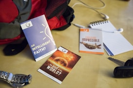A collection of items is arranged on a light-colored surface, including a red and gray backpack, a blue pamphlet titled 'An Intelligent Faith?', an orange pamphlet titled 'Evolution Impossible', and another with the title 'The End of the World?'. Additionally, there is a silver wristwatch, a set of car keys, a notepad with a pen, and white earphones.