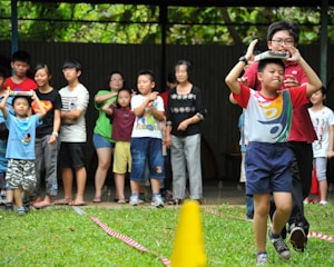 A group of children and adults are participating in an outdoor activity on grass. One child in the foreground is balancing an object on their head while walking, guided by an adult. The others stand in the background watching.