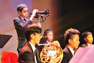 A group of young musicians dressed in formal attire are performing on stage. A trumpet player stands prominently in the foreground, playing their instrument with focus. Other musicians, including one holding a French horn, are seated and appear attentive, playing their parts. The setting is well-lit, creating a warm and vibrant atmosphere.