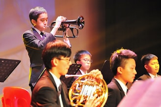 A group of young musicians dressed in formal attire are performing on stage. A trumpet player stands prominently in the foreground, playing their instrument with focus. Other musicians, including one holding a French horn, are seated and appear attentive, playing their parts. The setting is well-lit, creating a warm and vibrant atmosphere.