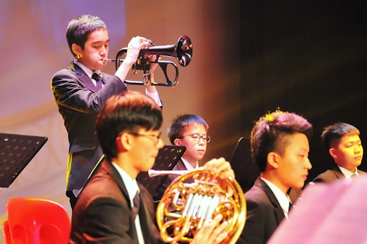 A group of young musicians dressed in formal attire are performing on stage. A trumpet player stands prominently in the foreground, playing their instrument with focus. Other musicians, including one holding a French horn, are seated and appear attentive, playing their parts. The setting is well-lit, creating a warm and vibrant atmosphere.