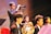 A group of young musicians dressed in formal attire are performing on stage. A trumpet player stands prominently in the foreground, playing their instrument with focus. Other musicians, including one holding a French horn, are seated and appear attentive, playing their parts. The setting is well-lit, creating a warm and vibrant atmosphere.