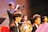 A group of young musicians dressed in formal attire are performing on stage. A trumpet player stands prominently in the foreground, playing their instrument with focus. Other musicians, including one holding a French horn, are seated and appear attentive, playing their parts. The setting is well-lit, creating a warm and vibrant atmosphere.