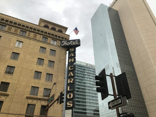A street view of a tall building with classic architecture featuring multiple windows and ornate details. A modern glass skyscraper is visible adjacent to it. The 'Hotel San Carlos' sign prominently stands in front of the buildings. An American flag is flying at the top of one of the structures. Traffic lights and a one-way street sign are in the foreground.