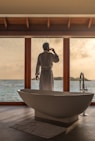 man standing in bathroom with bathtub next to body of water