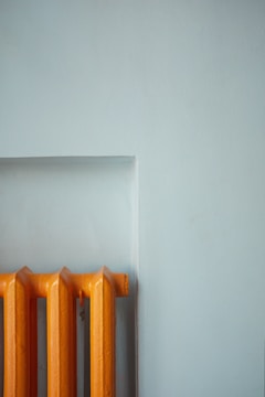 A vibrant orange radiator stands out against a muted light blue wall. The radiator's design features multiple evenly spaced vertical sections, showing a simple yet striking contrast in both color and form.