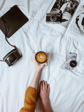 A cozy scene featuring a person with a warm mustard-colored sweater holding a latte with intricate latte art. Surrounding them on a white surface are a black leather purse, a vintage camera on an open magazine with black and white images, and a smartphone with a marble case and camera lens cap. The aesthetic suggests a casual and relaxed atmosphere.