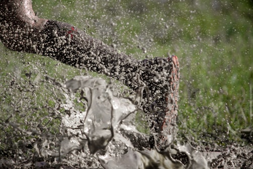A dynamic image captures a single leg, wearing a muddy shoe, sprinting through a splash of muddy water. Droplets are frozen mid-air, indicating motion and energy. The backdrop of blurred green suggests grass or a field environment.
