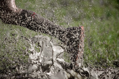 A dynamic image captures a single leg, wearing a muddy shoe, sprinting through a splash of muddy water. Droplets are frozen mid-air, indicating motion and energy. The backdrop of blurred green suggests grass or a field environment.