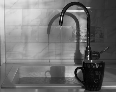 A monochrome image featuring a kitchen sink with a faucet running water into it. A ceramic mug with a coffee-themed design is placed on the edge of the sink. The background consists of a tiled backsplash with a subtle design, and soft shadows are cast on the wall.