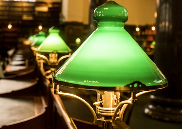 A row of traditional green glass library lamps is arranged on wooden desks, softly illuminating the surrounding area with a warm glow. The ambient lighting and classic design create a scholarly atmosphere, with bookshelves visible in the background.
