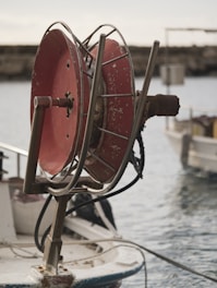 A large, metal hose reel with a worn red finish is mounted on the edge of a boat. The reel is rusted and scratches are visible, indicating extensive use. Behind the reel, a calm body of water and parts of other boats suggest a harbor or marina setting.