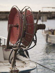 A large, metal hose reel with a worn red finish is mounted on the edge of a boat. The reel is rusted and scratches are visible, indicating extensive use. Behind the reel, a calm body of water and parts of other boats suggest a harbor or marina setting.