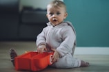 A young child wearing a light-colored knitted outfit is sitting on the floor, looking curiously forward. The child is holding a red, toy-like container with a logo on it. The background features a softly focused interior setting with muted colors.