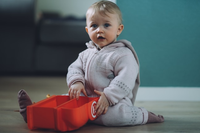 A young child wearing a light-colored knitted outfit is sitting on the floor, looking curiously forward. The child is holding a red, toy-like container with a logo on it. The background features a softly focused interior setting with muted colors.