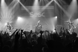 A musical performance features a band with three members on stage, surrounded by enthusiastic audience members raising their hands. Bright stage lights illuminate the scene, creating a dynamic and energetic atmosphere. The venue's signage is visible in the background.