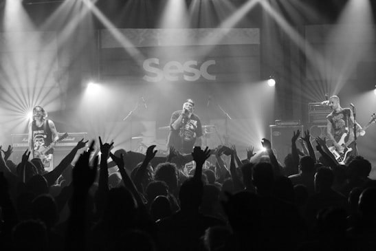 A musical performance features a band with three members on stage, surrounded by enthusiastic audience members raising their hands. Bright stage lights illuminate the scene, creating a dynamic and energetic atmosphere. The venue's signage is visible in the background.