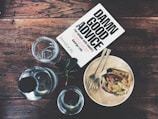 A wooden table holds a glass pitcher, two empty glasses, and a book titled 'Damn Good Advice' by George Lois. Beside the book is a wooden plate with a fork and knife resting on it, containing a pastry or sandwich with a visible tomato slice and melted cheese. A small sprig of greenery lies next to one of the glasses.