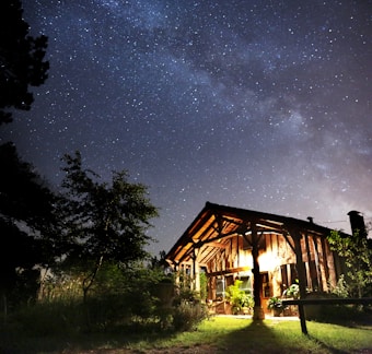 A small, rustic cabin illuminated by warm interior lights sits beneath a starry night sky. Surrounding the cabin are trees and lush greenery. The Milky Way is visible, adding a sense of wonder and tranquility to the scene.