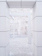 A modern architectural structure featuring clean lines and rectangular shapes dominates the scene. The image highlights an open space framed by large, white stone columns and walls, with visible block patterns. The flooring and textured stone elements add to the minimalist and sleek design.