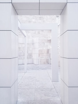 A modern architectural structure featuring clean lines and rectangular shapes dominates the scene. The image highlights an open space framed by large, white stone columns and walls, with visible block patterns. The flooring and textured stone elements add to the minimalist and sleek design.