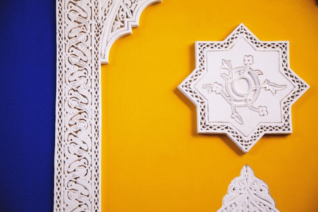 An ornate wall with a vibrant combination of blue and yellow paint. Intricate white stucco designs feature prominently, showcasing detailed patterns and a symmetrical star shape.