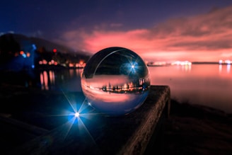 A crystal ball rests on a wooden surface, reflecting a vibrant and colorful sunset over a calm body of water. The sky is filled with dramatic clouds illuminated in shades of pink and orange, while lights from distant buildings line the horizon. A bright blue light creates a starburst effect near the foreground, adding a magical element to the scene.
