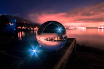 A crystal ball rests on a wooden surface, reflecting a vibrant and colorful sunset over a calm body of water. The sky is filled with dramatic clouds illuminated in shades of pink and orange, while lights from distant buildings line the horizon. A bright blue light creates a starburst effect near the foreground, adding a magical element to the scene.