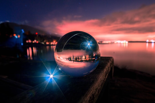 A crystal ball rests on a wooden surface, reflecting a vibrant and colorful sunset over a calm body of water. The sky is filled with dramatic clouds illuminated in shades of pink and orange, while lights from distant buildings line the horizon. A bright blue light creates a starburst effect near the foreground, adding a magical element to the scene.