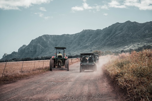 A rural dirt road stretches into the distance with a tractor and an off-road vehicle kicking up dust as they move away. The landscape is bordered by tall grass on one side and a fence on the other. Majestic, rugged mountains rise up in the background under a partly cloudy sky.