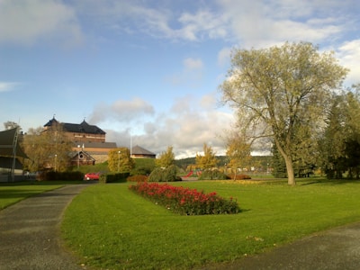 A well-maintained park with a pathway, neatly trimmed grass, and a large tree. In the background, an old brick building with a dark roof is visible, surrounded by smaller structures. A patch of red flowers adds a vibrant touch to the greenery.