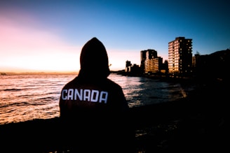 A person in a dark hoodie with 'CANADA' written on the back stands by the edge of a body of water, looking towards the ocean at sunset. Tall buildings line the shore, silhouetted against the colorful sky.