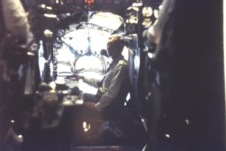 A person sits in a dimly lit cockpit with complex controls and instrumentation surrounding them. The setting appears to be inside an older aircraft or submarine, with a large window in front letting in some light.