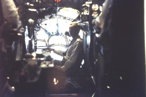 A person sits in a dimly lit cockpit with complex controls and instrumentation surrounding them. The setting appears to be inside an older aircraft or submarine, with a large window in front letting in some light.
